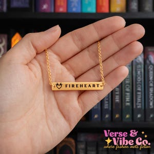 May include: A gold-colored necklace with a rectangular pendant that reads "FIREHEART" in black lettering. The pendant also features a heart and crown design. The necklace is held in a hand.