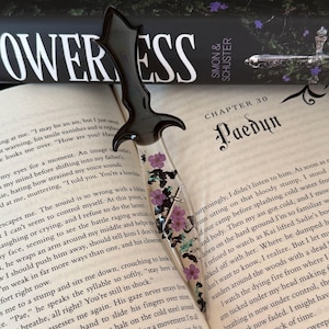 May include: A black dagger-shaped bookmark with a clear resin body, adorned with purple flowers and silver accents. The bookmark is resting on an open book with the title "POWERLESS" visible.