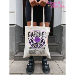 May include: A white tote bag with a purple and black graphic design featuring a rose and the text "Enemies to Lovers World Tour".