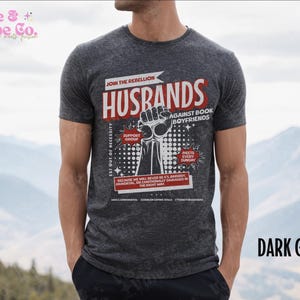 以下が含まれることがあります： ダークグレーのTシャツに、「Join the Rebellion: Husbands Against Book Boyfriends」というグラフィックプリントが施されています。グラフィックには、握りこぶしがあげられ、「Est out of necessity, Support Group, Meets Every Sunday, Because we will never be it's broody, hormonal, or emotionally damaged in the right way.」というテキストが書かれています。