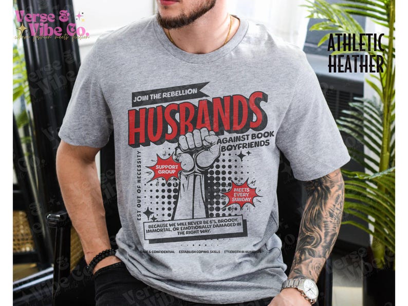 以下が含まれることがあります： コミック風のグラフィックがプリントされたグレーのTシャツ。"Husbands Join the Rebellion Against Book Boyfriends Est Out of Necessity Support Group Meets Every Sunday Because we will never be 6's, broody, immortal, or emotionally damaged in the right way."と書いてあります。
