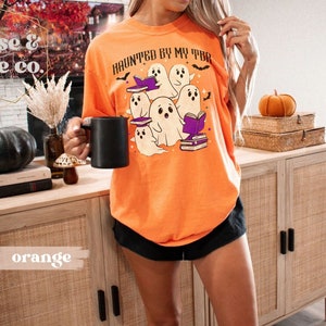 May include: An orange t-shirt featuring the phrase "HAUNTED BY MY TBR" and a graphic of cartoon ghosts reading books. The shirt is a loose fit, ideal for Halloween or book enthusiasts.