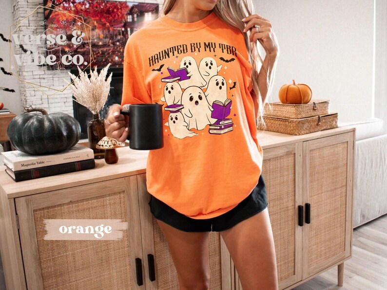 May include: An orange t-shirt featuring the phrase "HAUNTED BY MY TBR" and a graphic of cartoon ghosts reading books. The shirt is a loose fit, ideal for Halloween or book enthusiasts.