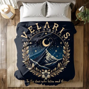 May include: A navy blue blanket with the text "VELARIS CITY OF STARLIGHT" in gold lettering. The design features a mountain range, a crescent moon, stars, and a floral wreath. The blanket also has the text "To the stars who listen and the..."