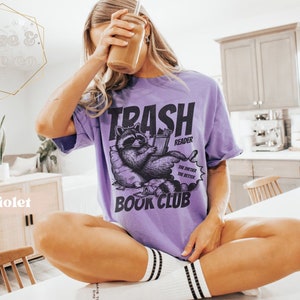 Puede incluir: Una camiseta morada con un gr&aacute;fico de un mapache leyendo un libro. El texto en la camiseta dice "Trash Reader The dirtier the better Book Club".