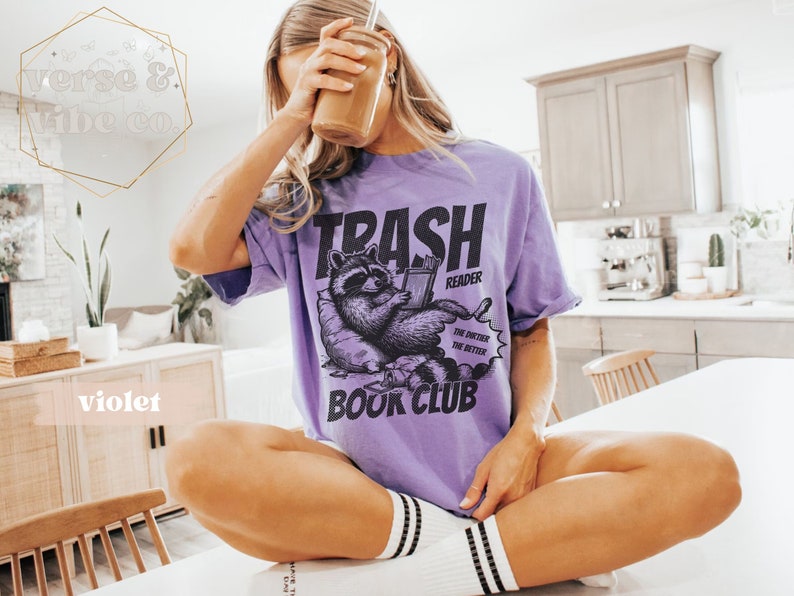 Puede incluir: Una camiseta morada con un gr&aacute;fico de un mapache leyendo un libro. El texto en la camiseta dice "Trash Reader The dirtier the better Book Club".