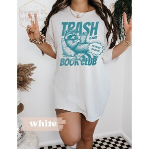 Puede incluir: Camiseta blanca con un gr&aacute;fico de un mapache leyendo un libro. El gr&aacute;fico dice "Trash Reader" y "The dirtier the better" con "Book Club" debajo.