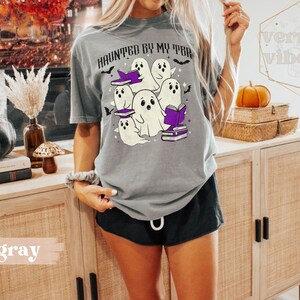May include: A grey t-shirt featuring the phrase "HAUNTED BY MY TBR" and a graphic of ghosts wearing witch hats and reading books. The design incorporates bats and purple details, ideal for Halloween or book enthusiasts.