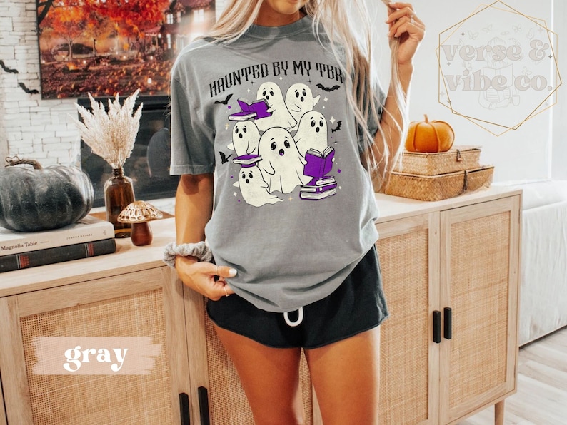 May include: A grey t-shirt featuring the phrase "HAUNTED BY MY TBR" and a graphic of ghosts wearing witch hats and reading books. The design incorporates bats and purple details, ideal for Halloween or book enthusiasts.