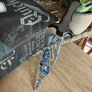 May include: A decorative dagger with a black handle and a clear blade filled with blue and pink glitter, butterflies, and small silver accents. The dagger rests on a wooden surface next to a book.