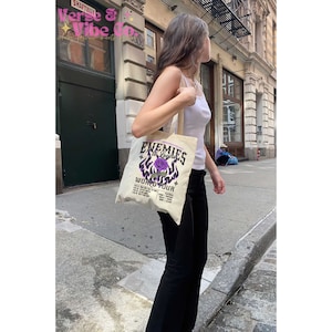 May include: A white canvas tote bag with a purple and black graphic design featuring a rose and flames. The text on the bag reads "Enemies World Tour".