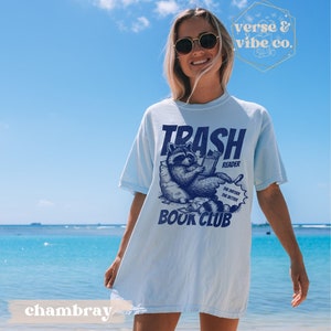 Puede incluir: Camiseta azul claro con un gr&aacute;fico de un mapache leyendo un libro. El texto en la camiseta dice "Trash Reader Book Club".