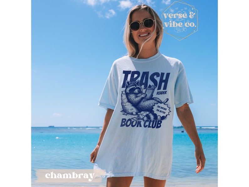 Puede incluir: Camiseta azul claro con un gr&aacute;fico de un mapache leyendo un libro. El texto en la camiseta dice "Trash Reader Book Club".
