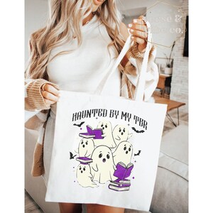May include: White tote bag featuring the phrase "HAUNTED BY MY TBR" and a graphic of cartoon ghosts reading books. The ghosts are white with purple books and accents, and black bats. The bag has white handles.