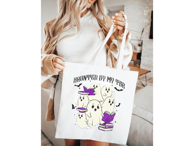 May include: White tote bag featuring the phrase "HAUNTED BY MY TBR" and a graphic of cartoon ghosts reading books. The ghosts are white with purple books and accents, and black bats. The bag has white handles.