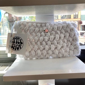 May include: White crocheted clutch bag with a small pink flower embellishment. The bag has a label that says "TYLE SHOP" with the text "handmade with love" underneath.
