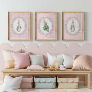 Puede incluir: Tres impresiones enmarcadas con ilustraciones de conejos en un estilo infantil clásico. Cada impresión tiene un fondo a rayas rosas y un marco ovalado decorativo. Los conejos visten chaquetas azules y verdes.