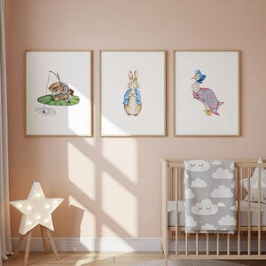 Könnte beinhalten: Drei gerahmte Aquarelldrucke mit Figuren aus Kinderbüchern. Einer zeigt ein Kaninchen in einer blauen Jacke, ein anderer eine Ente in einem rosa Kleid und der dritte ein Kaninchen beim Angeln. Die Drucke werden über einem Holzbettchen mit einer Wolkenmusterdecke angezeigt.