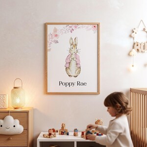 Puede incluir: Un cuadro enmarcado con una ilustración en acuarela de un conejo con una chaqueta rosa, con detalles florales. La impresión, etiquetada "Poppy Rae", está en un marco de madera clara. La obra de arte se exhibe en la habitación de un niño.