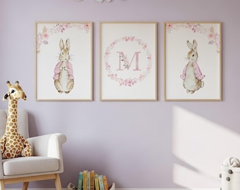 Könnte beinhalten: Drei gerahmte Kunstdrucke für das Kinderzimmer. Die Drucke zeigen ein Aquarell-Kaninchen mit rosa Pullover, einen Blumenkranz mit dem Buchstaben "M" und ein weiteres Kaninchen. Die Rahmen sind aus hellem Holz, der Hintergrund ist lavendelfarben.