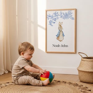 Puede incluir: Un marco de madera con una ilustración en acuarela de un conejo con una chaqueta azul, con detalles florales azules. El marco incluye el nombre "Noah John". Un bebé juega con una pelota colorida sobre una alfombra tejida.