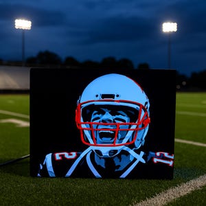 May include: A rectangular black canvas featuring a stylized portrait of a football player in a helmet with a red facemask. The player's face is illuminated in blue, with the numbers 12 and 11 visible on the jersey. The background is a football field.