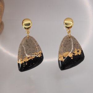 Puede incluir: Un par de pendientes colgantes dorados y negros con una base de resina gris y detalles de pan de oro.
