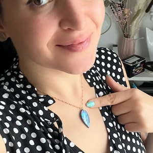 Peut inclure: Un collier en chaîne dorée avec un pendentif en forme de feuille bleu et iridescent. Le collier est porté par une personne avec une chemise à pois noirs et blancs.