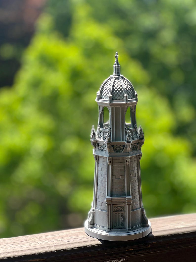 May include: A detailed, gray miniature lighthouse with a domed top and intricate architectural details. The lighthouse features a brick-like texture on its base and decorative elements around the top, set against a blurred green background.
