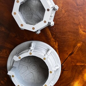 May include: Two light gray, open-top containers with intricate interior designs and gold-colored accents. One container is octagonal, the other is round. Both are set against a dark wood grain surface.