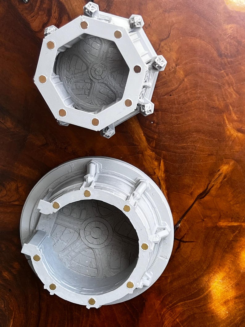 May include: Two light gray, open-top containers with intricate interior designs and gold-colored accents. One container is octagonal, the other is round. Both are set against a dark wood grain surface.