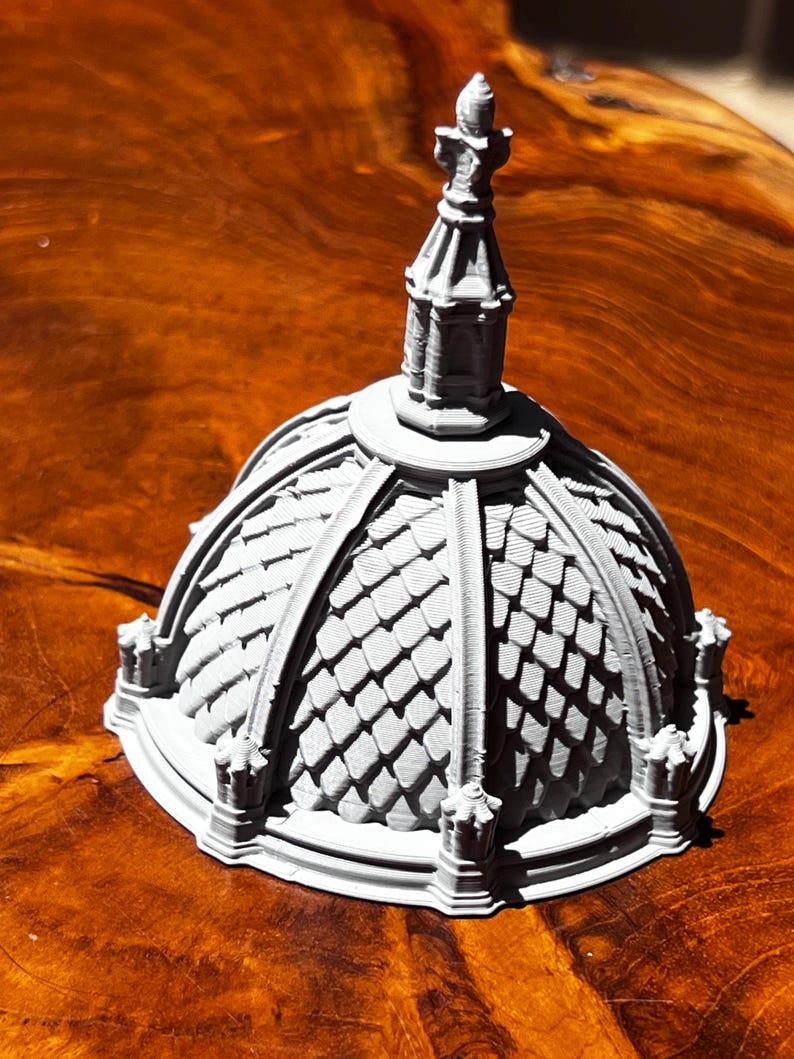 May include: A gray, 3D-printed architectural model of a domed structure with a detailed roof and a spire. The model features intricate detailing and a textured surface, showcasing a miniature replica of a building.