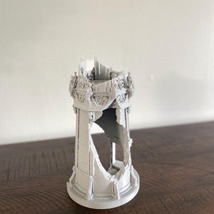 May include: A gray 3D-printed model of a ruined tower. The tower has intricate architectural details, with a large section missing, revealing the interior. The model sits on a dark wooden surface.