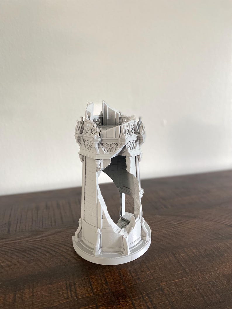 May include: A gray 3D-printed model of a ruined tower. The tower has intricate architectural details, with a large section missing, revealing the interior. The model sits on a dark wooden surface.
