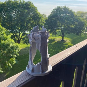 May include: A gray, 3D-printed model of a ruined tower, featuring intricate architectural details. The tower has a circular base and is partially destroyed, with visible openings. The model is set against a backdrop of green trees and a blue sky.