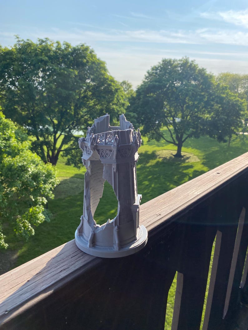 May include: A gray, 3D-printed model of a ruined tower, featuring intricate architectural details. The tower has a circular base and is partially destroyed, with visible openings. The model is set against a backdrop of green trees and a blue sky.