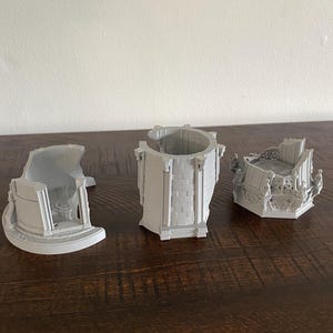 May include: Three gray 3D-printed architectural models of a building, each with a different section. The models are placed on a dark brown wooden surface. The models appear to be a tower and two other building sections.