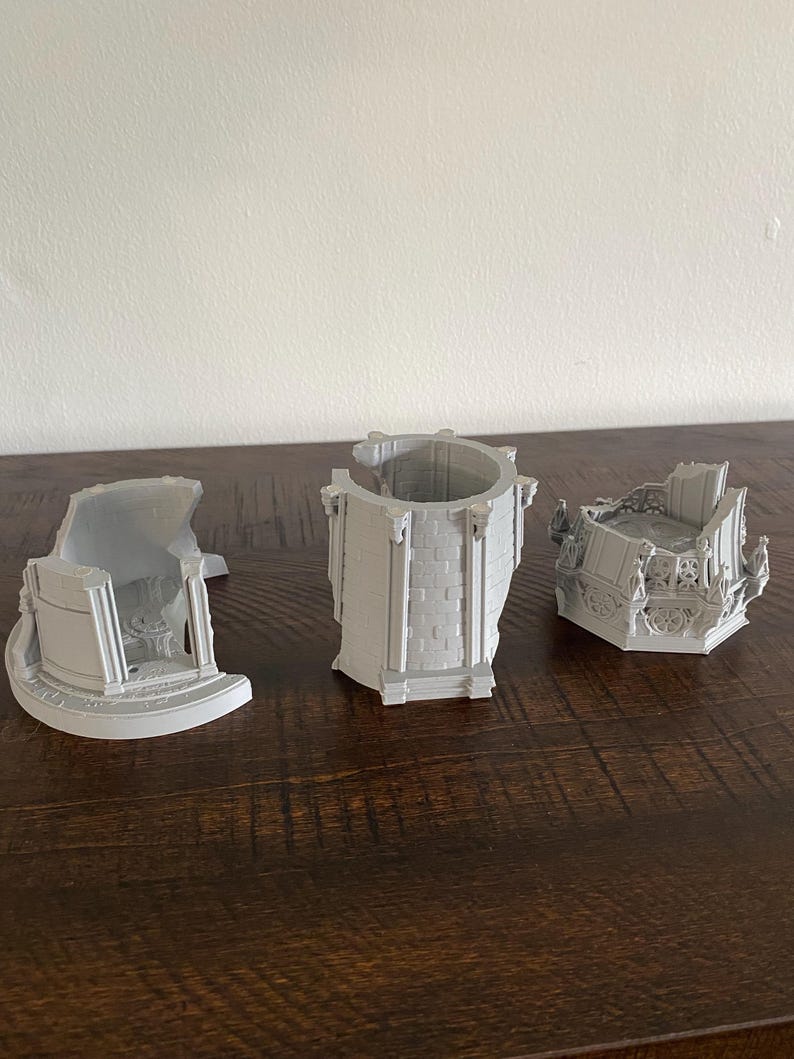 May include: Three gray 3D-printed architectural models of a building, each with a different section. The models are placed on a dark brown wooden surface. The models appear to be a tower and two other building sections.