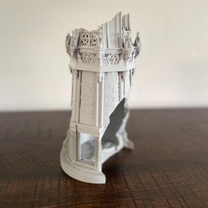 May include: A gray, 3D-printed model of a ruined castle tower. The tower features detailed brickwork, decorative elements, and a partially collapsed structure, showcasing architectural detail. The model is displayed on a dark wooden surface.