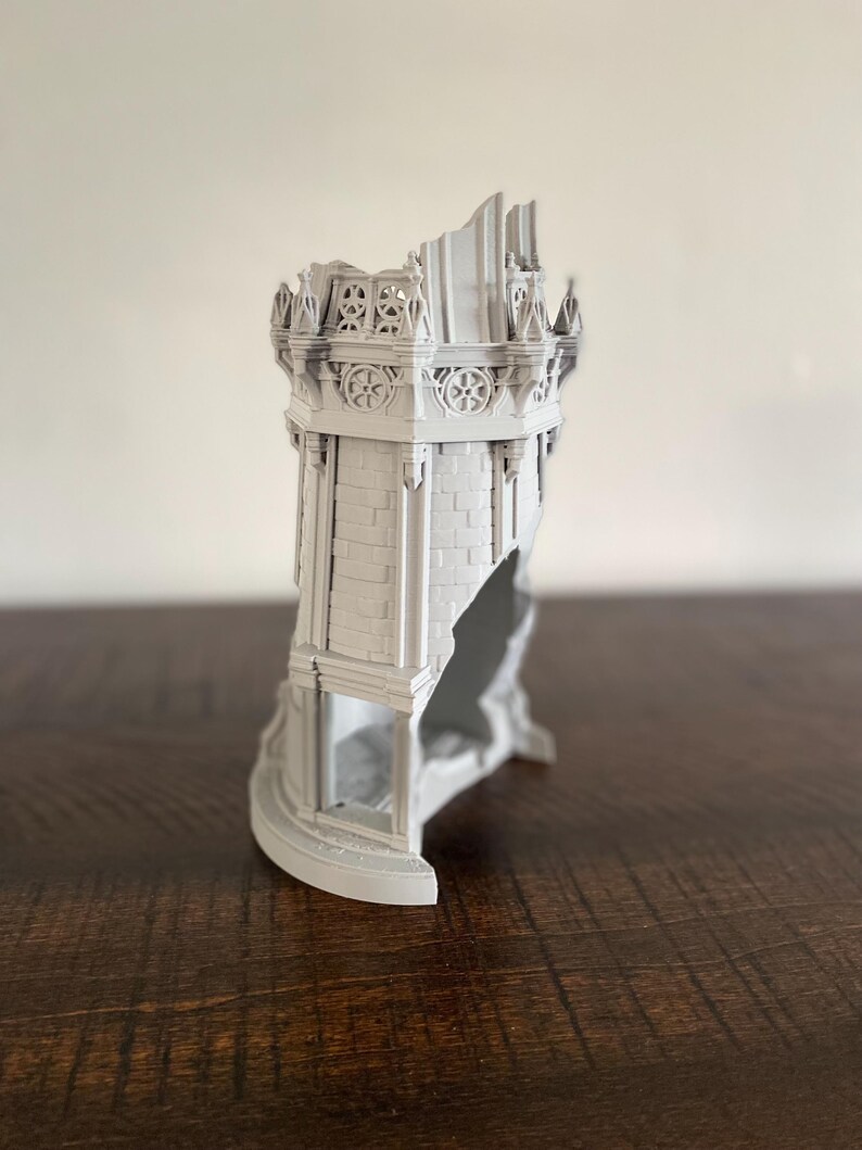 May include: A gray, 3D-printed model of a ruined castle tower. The tower features detailed brickwork, decorative elements, and a partially collapsed structure, showcasing architectural detail. The model is displayed on a dark wooden surface.