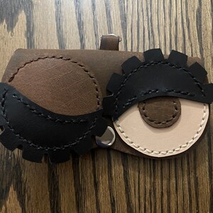 May include: A brown leather eyeglass case with two large, black leather eyes with brown leather pupils. The case has a stitched design and a metal clasp.
