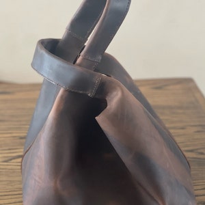 Könnte beinhalten: Eine braune Ledertasche mit einem einzigen, dicken Henkel. Die Tasche ist gefaltet und hat einen leicht abgenutzten Look.