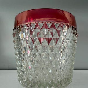 May include: A clear glass jar with a diamond pattern and a red rim. The jar is cylindrical and has a textured surface. The red rim adds a pop of color to the otherwise transparent container. This decorative jar is suitable for storage or display.