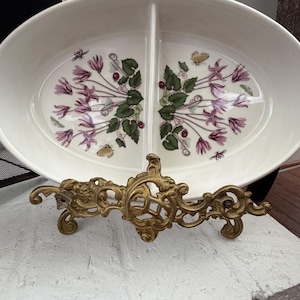 May include: An oval, divided serving dish with a floral design of pink flowers, green leaves, and small insects. The dish is supported by an ornate, gold-colored metal stand. The dish is white.