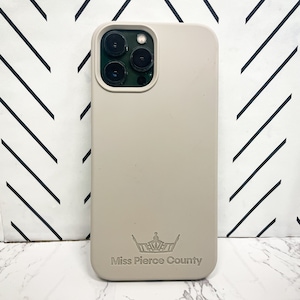 May include: A light beige phone case with a crown logo and the text "Miss Pierce County" is displayed. The case has a smooth finish and a camera cutout. The background is a black and white chevron pattern.