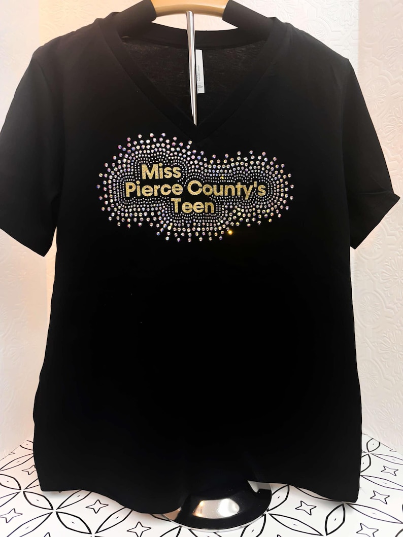 Puede incluir: Camiseta negra de cuello en V con un dise&ntilde;o de purpurina dorada que dice "Miss Pierce County's Teen".