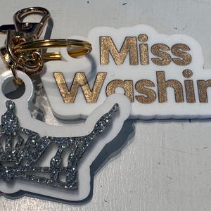 May include: A white keychain with a gold-colored clasp and tassel. The keychain features a crown-shaped charm with silver glitter and a rectangular charm with the text "Miss Washington" in gold lettering.