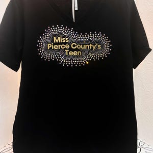 Puede incluir: Camiseta negra de cuello en V con un dise&ntilde;o de purpurina dorada que dice "Miss Pierce County's Teen".