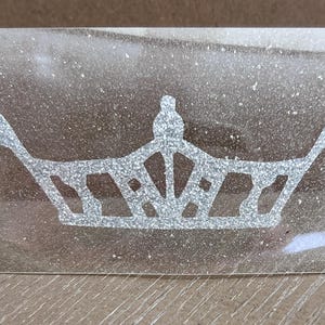May include: A silver glitter crown with a pointed top and a decorative design. The crown is on a clear background.
