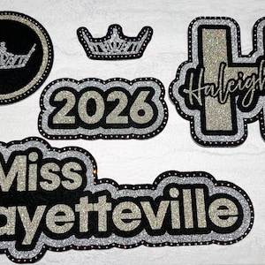 May include: A collection of black and silver glitter embellishments, including a crown, a circle with a crown, the number "2026", the name "Haleigh Jo", and the words "Miss Fayetteville". These items are likely for crafts or decorations.